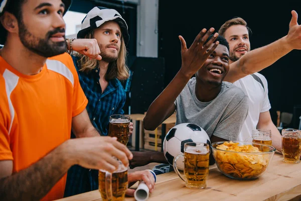 Sonriente grupo multicultural de fanáticos del fútbol masculino haciendo gestos con las manos y viendo el partido de fútbol en el bar - foto de stock