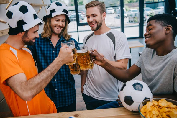 Sonriente grupo multicultural de aficionados al fútbol masculino tintineo vasos de cerveza durante el reloj del partido de fútbol en el bar - foto de stock
