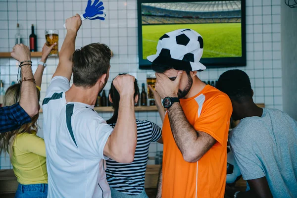 Jovem frustrado em chapéu de bola de futebol e camiseta de ventilador segurando olhos cobrindo palma da mão, enquanto seus amigos comemorando e fazendo gestos sim durante o relógio de jogo de futebol na tela da TV no bar — Fotografia de Stock