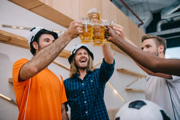 Low angle view of football fans in soccer ball hats celebrating victory and clinking beer glasses at bar — Stock Photo