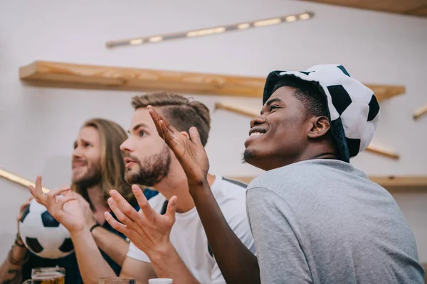 Side view of multicultural group of male football fans gesturing by hands and watching soccer match at bar — Stock Photo