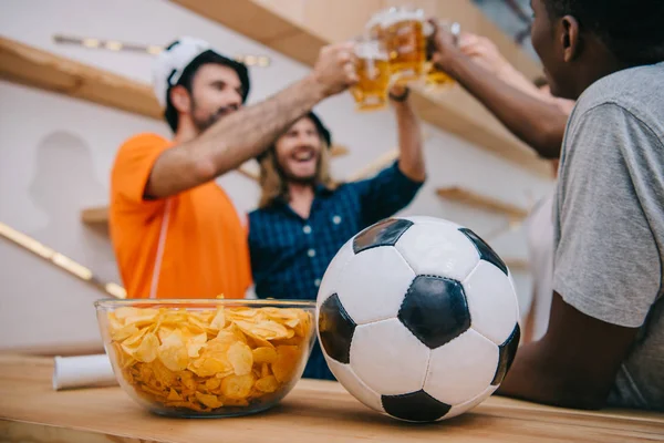 Vista de primer plano de la pelota de fútbol y el tazón con fichas con el grupo de aficionados al fútbol celebrando y tintineo por vasos de cerveza detrás en el bar - foto de stock