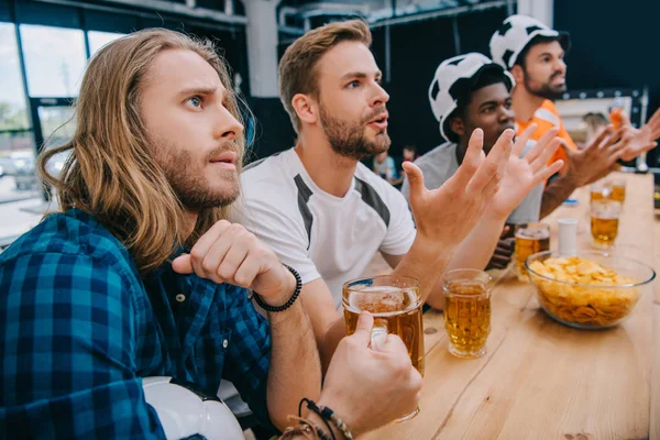 Molesto grupo multicultural de fanáticos del fútbol masculino en sombreros de fútbol bebiendo cerveza y viendo el partido de fútbol en el bar - foto de stock