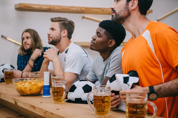 Multicultural group of male football fans with soccer ball hats, fan horn, chips and beer watching soccer match at bar — Stock Photo