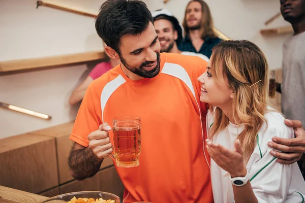 Lächelnder Mann mit Bier umarmt Freundin und ihre multikulturellen Freunde beim Fußballspiel in der Bar — Stockfoto