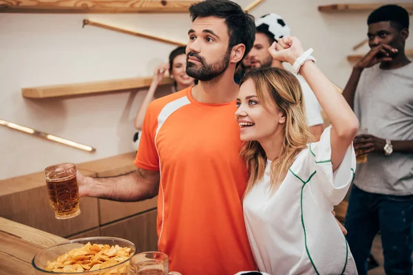 Pareja joven con amigos multiculturales celebrando y viendo el partido de fútbol en el bar - foto de stock