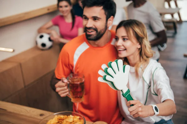 Smiling young couple with beer and hand clappers watching soccer match at bar — Stock Photo