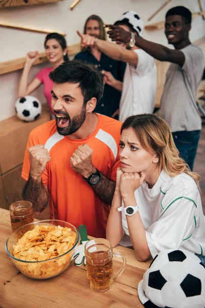 Aficionados al fútbol multicultural emocional señalando con los dedos y viendo el partido de fútbol en el bar con patatas fritas y vasos de cerveza - foto de stock