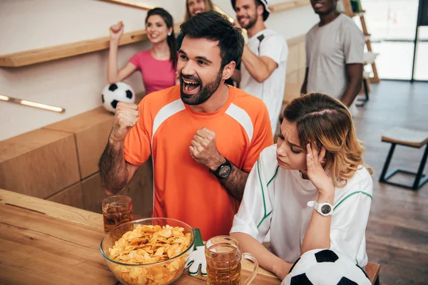 Sonriente joven en naranja ventilador camiseta celebrando y haciendo sí gestos mientras que su novia molesta sentado con la mano en el templo en el bar con patatas fritas y cerveza - foto de stock