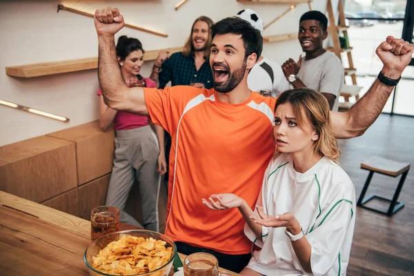 Smiling young man in orange fan t-shirt celebrating with wide raised arms and his upset girlfriend in fan t-shirt sitting and gesturing by hands at bar with chips and beer — Stock Photo