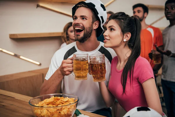 Hombre emocionado en el sombrero de pelota de fútbol tintineo vasos de cerveza con la novia, mientras que sus amigos multiculturales viendo partido de fútbol en el bar - foto de stock