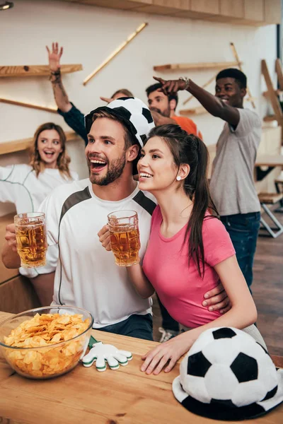 Homem feliz em chapéu de bola de futebol abraçando namorada e segurando cerveja, enquanto seus amigos comemorando atrás e gesticulando por mãos durante o relógio de jogo de futebol no bar — Fotografia de Stock
