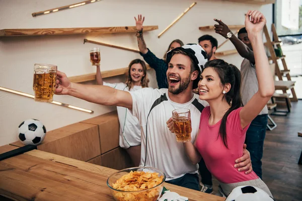Vue grand angle de l'homme heureux dans le chapeau de football embrassant petite amie et tenant de la bière tandis que leurs amis célébrant derrière et regarder le match de football au bar — Photo de stock