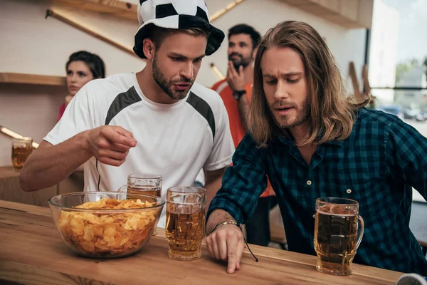 Joven con sombrero de fútbol haciendo gestos y hablando con su amigo sentado en el bar con cerveza y patatas fritas durante el reloj del partido de fútbol - foto de stock