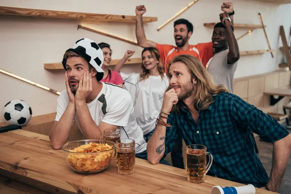 Excited group of multicultural friends celebrating and watching soccer match at bar with beer and chips — Stock Photo