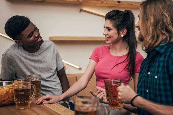 Young african american man talking to friends and sitting at bar counter with chips and beer — Stock Photo