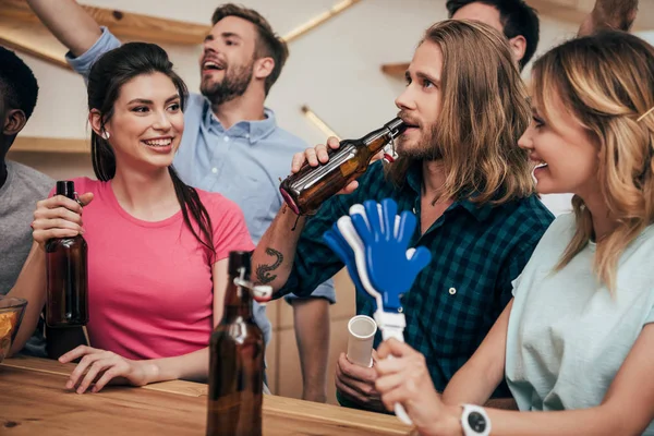 Улыбающиеся мультикультурные друзья, празднующие с фанатскими рогами и хлопающими руками во время просмотра футбольного матча в баре — стоковое фото