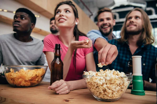 Низкоугольный вид на многоэтническую группу друзей, смотрящих футбольный матч в баре с чипсами, кукурузой, фанатским рожком и пивными бутылками — стоковое фото