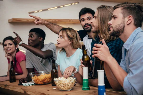 Two men pointing by hands to friends sitting at bar with fan horns, popcorn, chips and beer bottles during watch of football match — Stock Photo