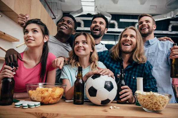 Smiling multiethnic group of friends watching football match at bar with chips, popcorn, beer and soccer ball — Stock Photo