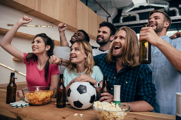 Feliz grupo multiétnico de amigos celebrando, haciendo gestos de sí y viendo el partido de fútbol en el bar con patatas fritas, cerveza y pelota de fútbol - foto de stock