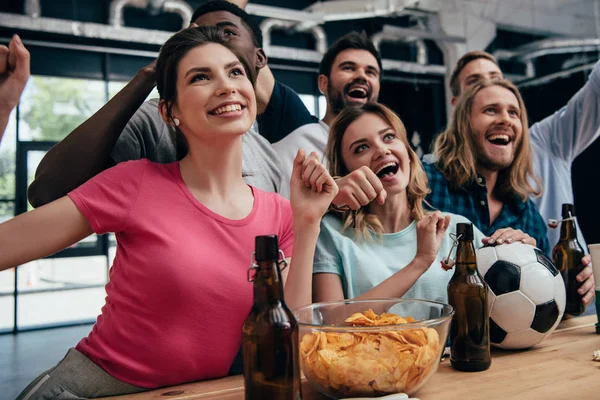 Feliz grupo multiétnico de amigos celebrando y viendo el partido de fútbol en el bar con patatas fritas, cerveza y pelota de fútbol - foto de stock