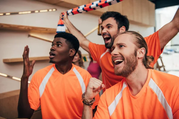 Excited multicultural male football fans in orange t-shirts celebrating and gesturing during watch of soccer match at bar — Stock Photo