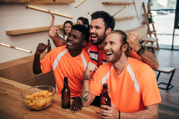 Amis souriants multiculturels en t-shirts orange faire des gestes oui et assis au bar avec de la bière et des frites pendant regarder le match de football — Photo de stock