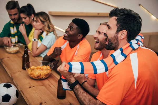 Vista lateral de felices jóvenes amigos multiculturales en naranja ventilador camisetas celebrando gol en el partido de fútbol, mientras que sus amigos sentados disgustados en el otro lado del mostrador de bar - foto de stock
