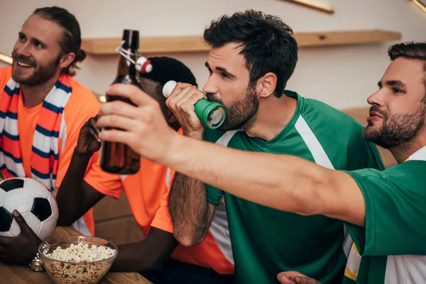 Grupo de fanáticos del fútbol multicultural en camisetas naranjas y verdes viendo el partido de fútbol en el bar - foto de stock