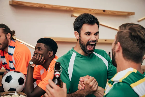 Excited football fans in green t-shirts celebrating victory while their upset friends in different t-shirts sitting behind with ball during watch of soccer match at bar — Stock Photo