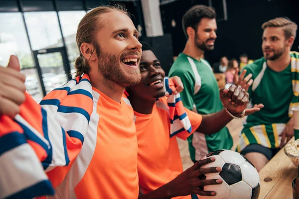 Excited multicultural football fans in orange t-shirts and scarf celebrating victory with ball while their upset friends in different t-shirts sitting behind during watch of soccer match at bar — Stock Photo