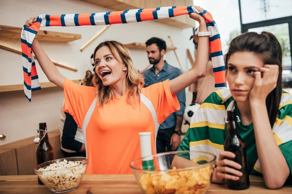 Excited young woman in orange fan t-shirt celebrating and holding fan scarf over head while her upset female friend in green t-shirt sitting with beer bottle during watch of soccer match — Stock Photo