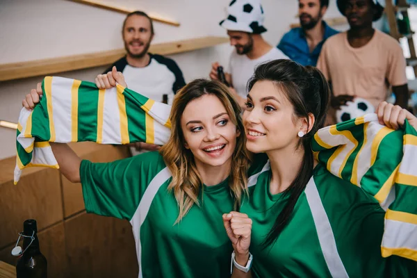 Femmes en t-shirts vert ventilateur tenant écharpe ventilateur et leurs amis masculins debout derrière pendant la montre du match de football au bar — Photo de stock