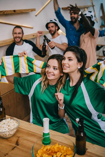 Vista de ángulo alto de las mujeres en verde ventilador camisetas que sostienen la bufanda del ventilador y sus amigos masculinos de pie durante el reloj del partido de fútbol en el bar - foto de stock