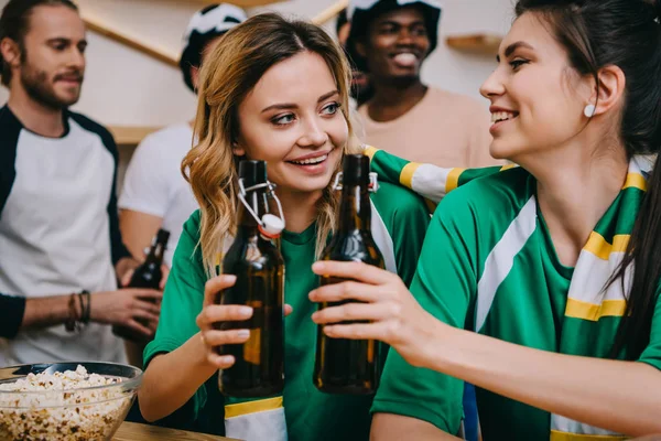 Sonrientes mujeres jóvenes tintineo botellas de cerveza y sus amigos masculinos de pie durante el reloj de fútbol partido en el bar - foto de stock