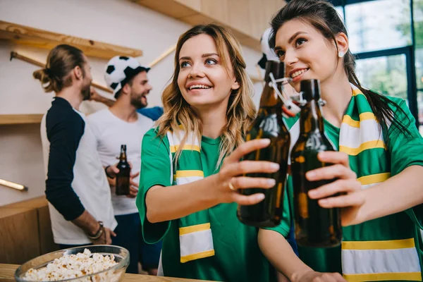 Feliz jóvenes mujeres tintineo botellas de cerveza y sus amigos masculinos de pie detrás durante el reloj de fútbol partido en el bar - foto de stock