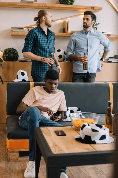 Jeune homme afro-américain utilisant un smartphone sur le canapé tandis que ses amis parlent avec des bouteilles de bière derrière à la maison — Photo de stock