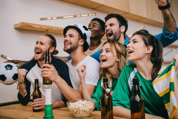 Amigos multiculturales emocionados con botellas de cerveza haciendo gestos de sí durante el reloj del partido de fútbol en el bar - foto de stock