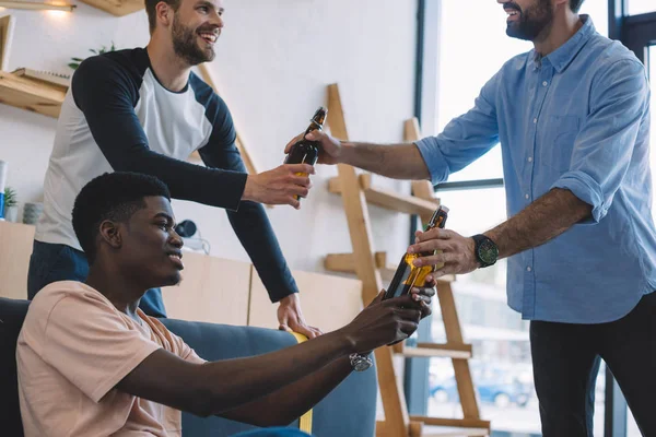 Cropped shot of man giving beer bottles to smiling multiethnic friends at home — Stock Photo