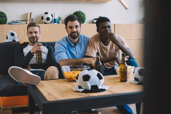 Jovens amigos multiculturais assistindo jogo de futebol perto da mesa com batatas fritas, pipocas e chapéus de bola em casa — Stock Photo