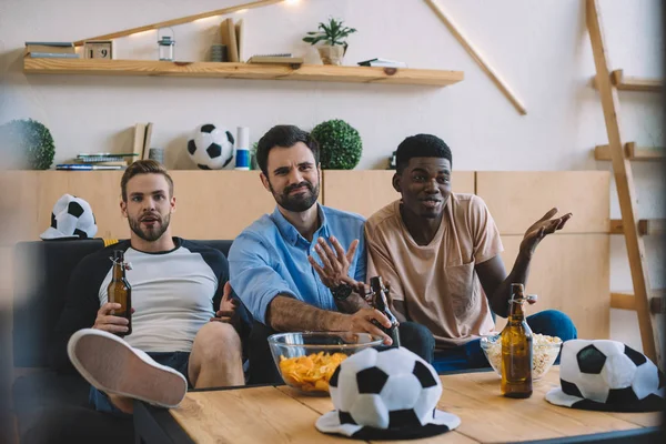 Molestar a jóvenes amigos multiculturales sacudiendo botellas de cerveza y celebrando durante el reloj del partido de fútbol en casa - foto de stock