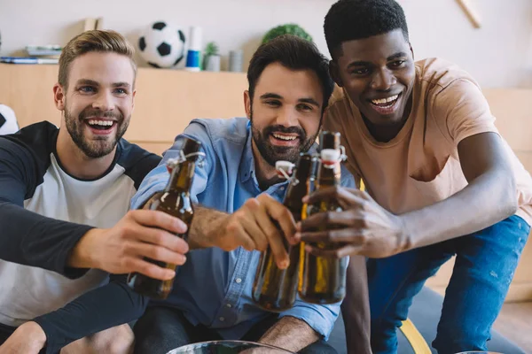 Jovens amigos multiculturais felizes batendo garrafas de cerveja e celebrando durante o relógio de jogo de futebol em casa — Fotografia de Stock