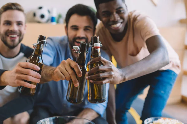 Emocionados jóvenes amigos multiculturales tintinear botellas de cerveza y celebrar durante el reloj del partido de fútbol en casa - foto de stock