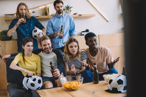 Sorrindo amigos multiculturais garrafas de cerveja sentado no sofá perto da mesa com pipocas e batatas fritas assistindo jogo de futebol em casa — Fotografia de Stock