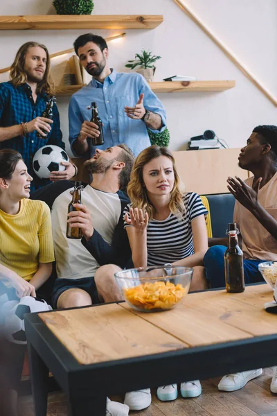 Amigos multiculturales emocionales botellas de cerveza sentados en el sofá cerca de la mesa con palomitas de maíz y patatas fritas viendo el partido de fútbol en casa - foto de stock