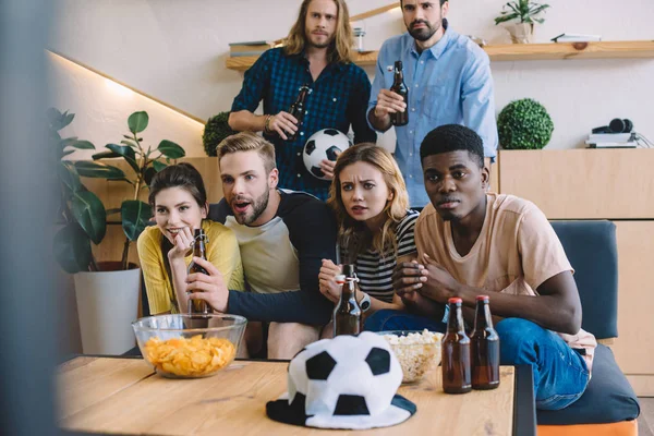 Amigos multiculturales botellas de cerveza sentados en el sofá cerca de la mesa con palomitas de maíz y patatas fritas viendo el partido de fútbol en casa - foto de stock