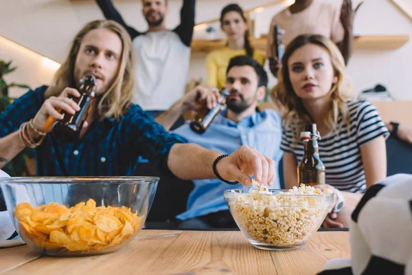 Young man drinking beer and taking popcorn from bowl during watch of football match with friends at home — Stock Photo