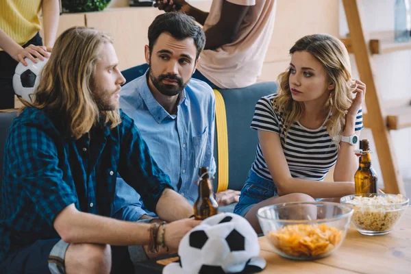 Jóvenes fanáticos del fútbol sentados en el sofá con botellas de cerveza y hablando entre sí cerca de la mesa con patatas fritas y palomitas de maíz - foto de stock