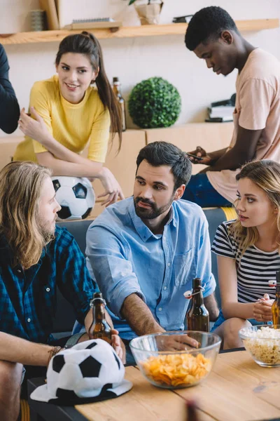 Grupo fãs de futebol multicultural sentados no sofá e conversando uns com os outros perto da mesa com cerveja, batatas fritas e pipocas — Fotografia de Stock